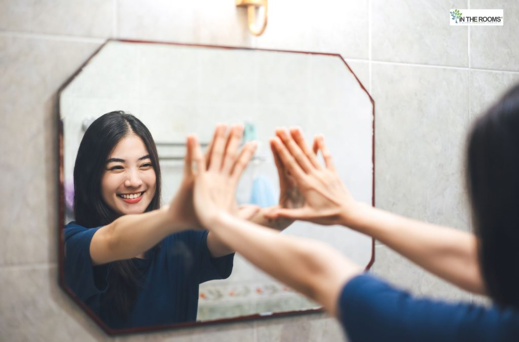 Woman smiling and touching hands with her reflection in the mirror, practicing self-talk
