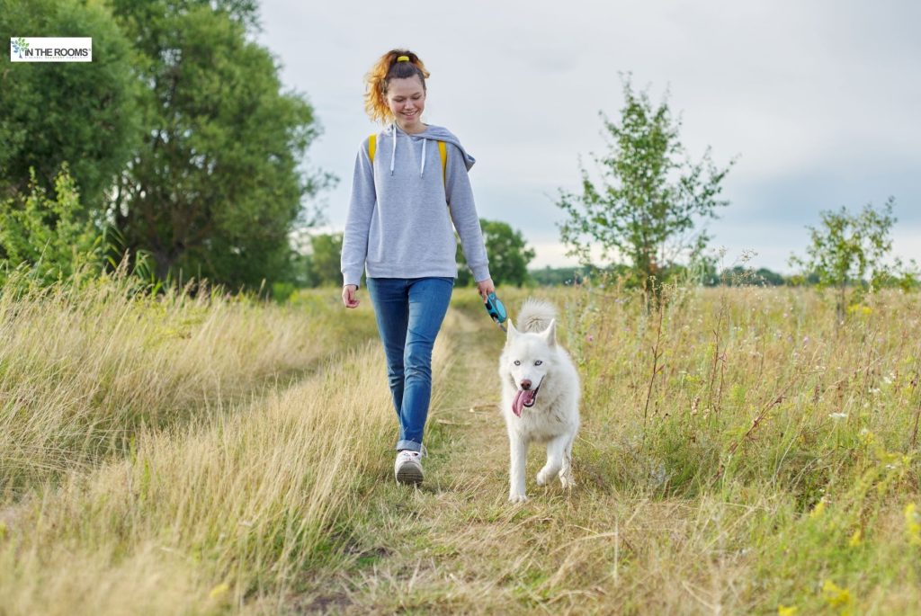 Smiling woman walking with a white dog on a grassy path surrounded by trees and wildflowers.