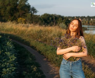 Woman standing outdoors on a dirt path, holding a bouquet of wildflowers with eyes closed, embracing peace and self-love.