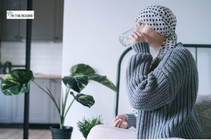 Woman wearing a polka-dotted headscarf and gray sweater sits indoors, drinking water, reflecting on her recovery journey.