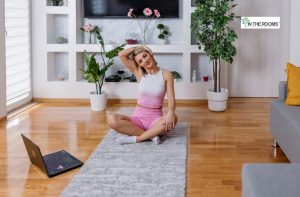 Woman in workout clothes sitting on a yoga mat indoors, stretching her neck while following a morning routine with a laptop nearby.