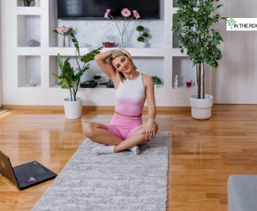 Woman in workout clothes sitting on a yoga mat indoors, stretching her neck while following a morning routine with a laptop nearby.