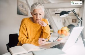 Mature woman with short gray hair wearing a mustard yellow sweater, sitting at a desk with a laptop and notebook, appearing thoughtful as she studies or reflects.