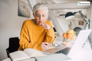 Mature woman with short gray hair wearing a mustard yellow sweater, sitting at a desk with a laptop and notebook, appearing thoughtful as she studies or reflects.