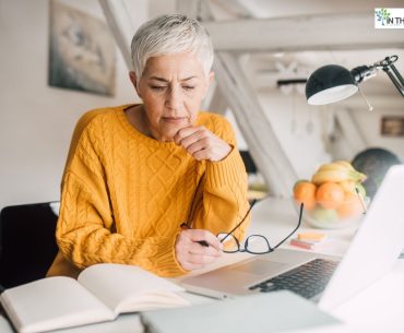 Mature woman with short gray hair wearing a mustard yellow sweater, sitting at a desk with a laptop and notebook, appearing thoughtful as she studies or reflects.
