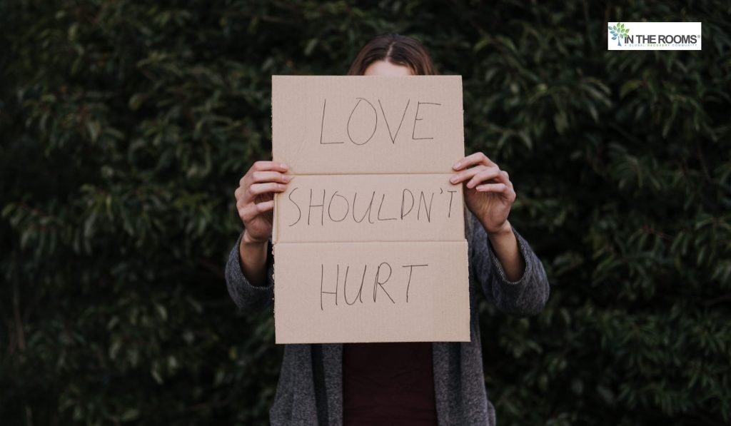 Person standing outdoors holding a cardboard sign that reads “LOVE SHOULDN’T HURT,” symbolizing emotional recovery and the courage to speak out against abuse.