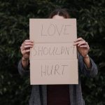 Person standing outdoors holding a cardboard sign that reads “LOVE SHOULDN’T HURT,” symbolizing emotional recovery and the courage to speak out against abuse.