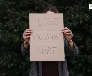 Person standing outdoors holding a cardboard sign that reads “LOVE SHOULDN’T HURT,” symbolizing emotional recovery and the courage to speak out against abuse.