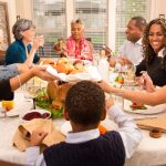 A diverse family and friends group sitting around a Thanksgiving dinner table, smiling, sharing food, and enjoying a warm holiday meal together.