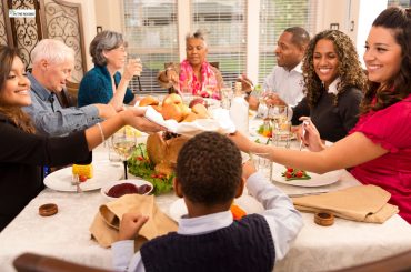 A diverse family and friends group sitting around a Thanksgiving dinner table, smiling, sharing food, and enjoying a warm holiday meal together.