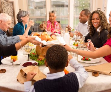 A diverse family and friends group sitting around a Thanksgiving dinner table, smiling, sharing food, and enjoying a warm holiday meal together.