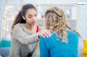 A young woman gently placing her hand on another person’s shoulder, offering comfort and support during an emotional moment.