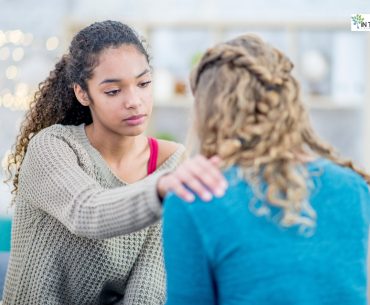 A young woman gently placing her hand on another person’s shoulder, offering comfort and support during an emotional moment.
