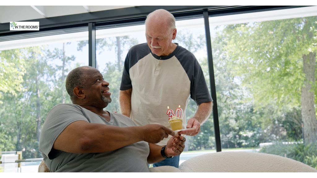 Two men smiling as one hands the other a cupcake with “30” candles, celebrating a sobriety milestone in a bright, peaceful room.