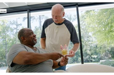 Two men smiling as one hands the other a cupcake with “30” candles, celebrating a sobriety milestone in a bright, peaceful room.
