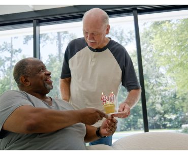 Two men smiling as one hands the other a cupcake with “30” candles, celebrating a sobriety milestone in a bright, peaceful room.