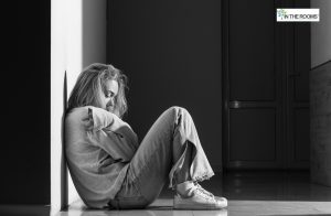 A woman sitting on the floor against a wall, hugging her knees and looking down, expressing sadness and vulnerability after a difficult moment.