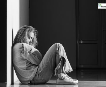 A woman sitting on the floor against a wall, hugging her knees and looking down, expressing sadness and vulnerability after a difficult moment.