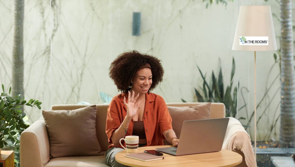 Woman sitting on a cozy sofa, smiling and waving at her laptop during a virtual conversation, symbolizing connection and personal growth.