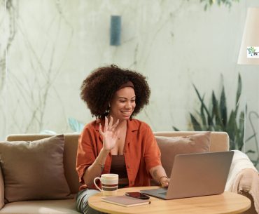 Woman sitting on a cozy sofa, smiling and waving at her laptop during a virtual conversation, symbolizing connection and personal growth.