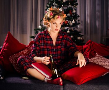 A woman sitting on a couch in holiday pajamas near a Christmas tree, holding a wine bottle and looking distressed, symbolizing emotional triggers and cravings during the holiday season.