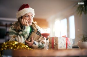 A woman wearing a Santa hat sits alone at a table with holiday gifts and a glass of wine nearby, looking thoughtful and isolated during the holiday season.