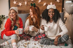 Three women in cozy holiday outfits laughing and enjoying warm drinks together at a festive gathering, symbolizing sober celebration and supportive friendships.