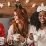 Three women in cozy holiday outfits laughing and enjoying warm drinks together at a festive gathering, symbolizing sober celebration and supportive friendships.