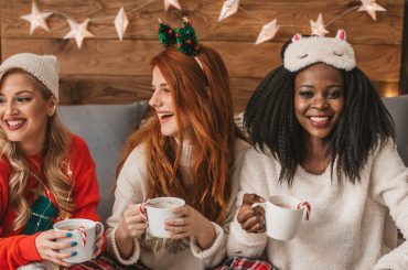 Three women in cozy holiday outfits laughing and enjoying warm drinks together at a festive gathering, symbolizing sober celebration and supportive friendships.