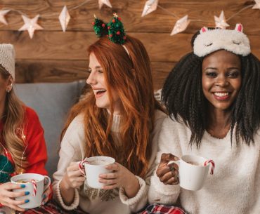 Three women in cozy holiday outfits laughing and enjoying warm drinks together at a festive gathering, symbolizing sober celebration and supportive friendships.