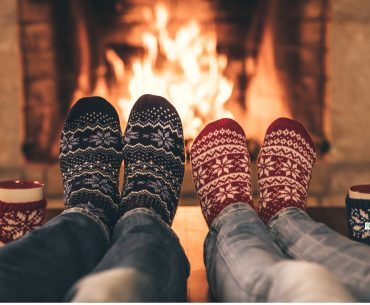 Two people in festive socks relaxing by a warm fireplace with mugs beside them, symbolizing calm, comfort, and a safe space during the holiday season.
