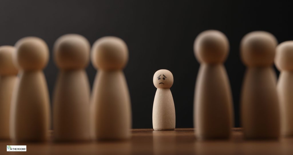 A small wooden figure standing alone between two blurred groups of wooden figures, symbolizing loneliness and isolation during recovery.