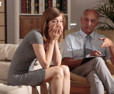 A woman sitting on a couch with her hands covering her mouth during a therapy session, while a therapist listens and takes notes.
